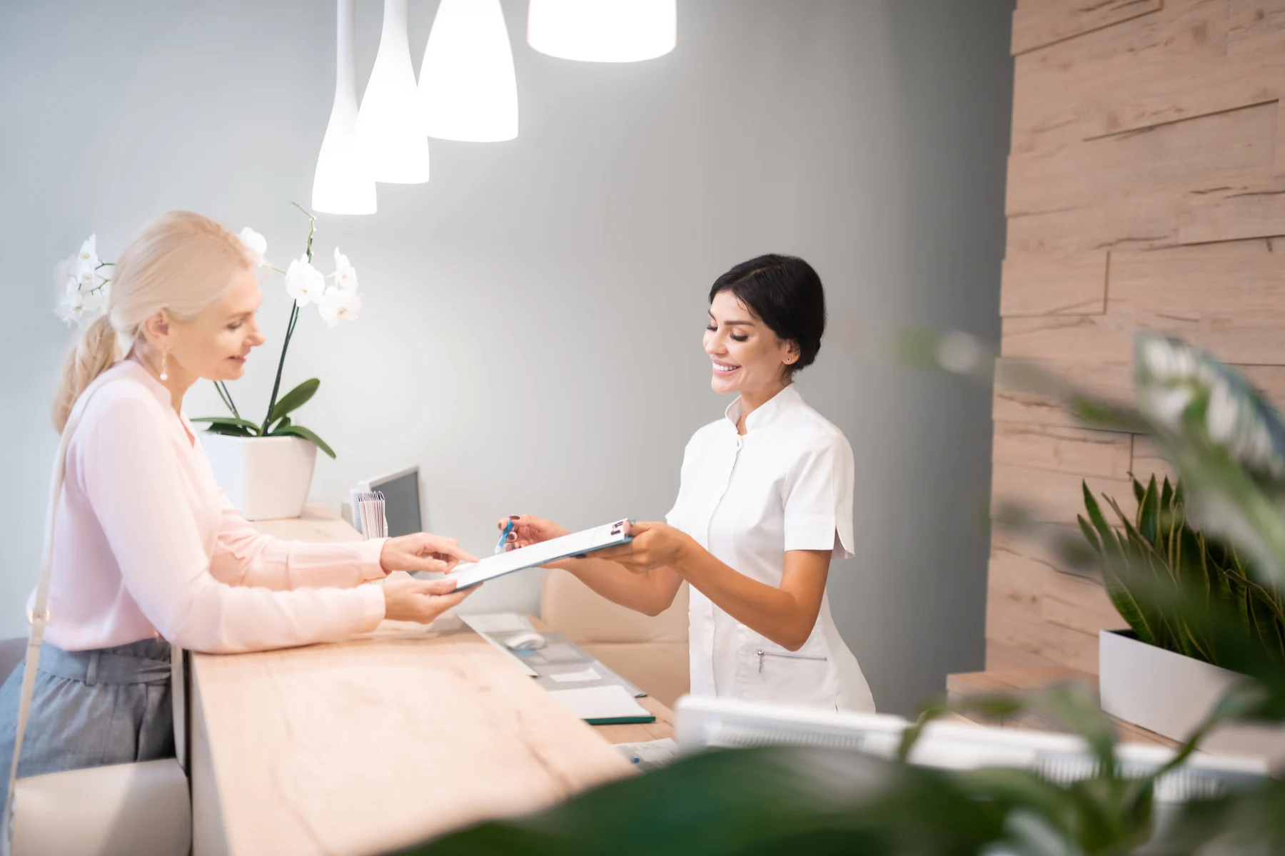 Profesional de una clínica atendiendo a una mujer en una recepción luminosa con madera y plantas.